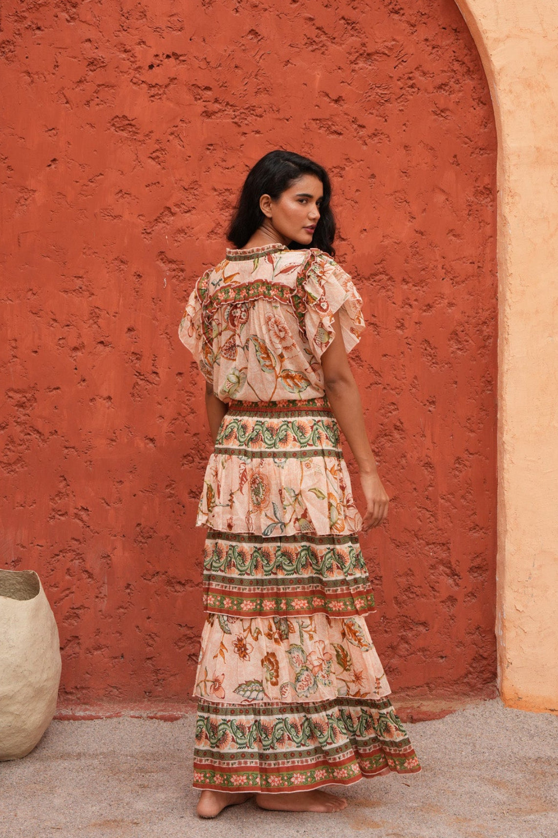 A woman stands in front of a textured terracotta archway wearing a long, tiered bohemian maxi dress with a beige base and colorful floral patterns. The scene is set in a warm, rustic outdoor environment with earthy clay pots on the ground.
