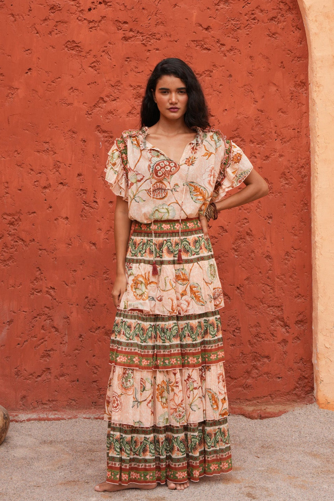 A woman stands in front of a textured terracotta archway wearing a bohemian-style maxi dress with a cream base and colorful floral patterns. The dress features short flutter sleeves, a V-neckline, and a tiered skirt with detailed borders.