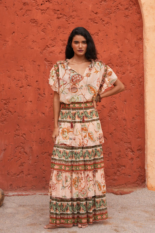 A woman stands in front of a textured terracotta archway wearing a bohemian-style maxi dress with a cream base and colorful floral patterns. The dress features short flutter sleeves, a V-neckline, and a tiered skirt with detailed borders.