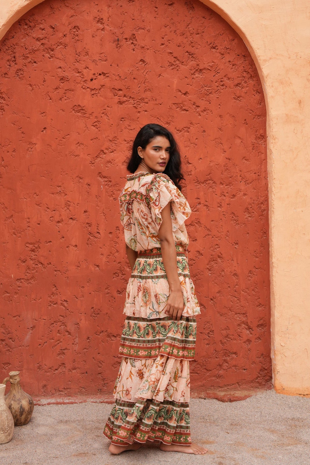 A woman stands in front of a large terracotta archway wearing a long, tiered bohemian maxi dress with a cream base and green, orange, and red floral patterns. The background consists of a textured orange wall and a few rustic clay pots on the left.