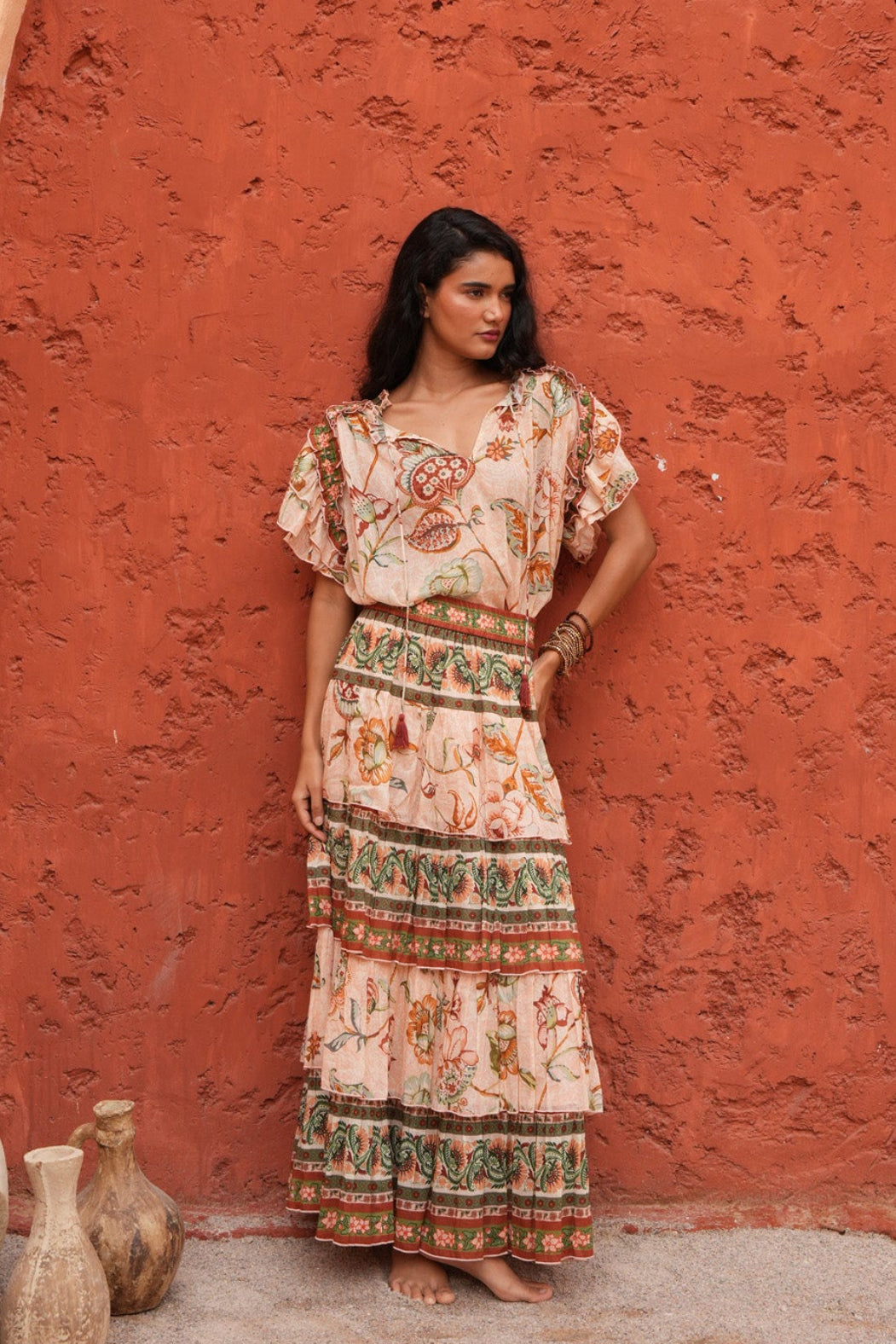 A woman stands barefoot in front of a textured terracotta archway, wearing a bohemian-style maxi dress with a cream base and colorful floral patterns. The scene is set against a warm, peach-colored stucco wall with decorative clay pots on the ground.
