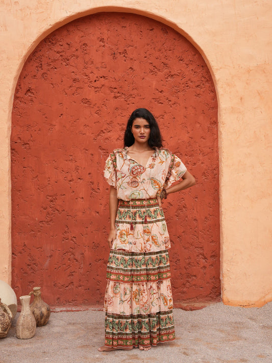 A woman stands in front of a textured terracotta archway wearing a bohemian-style maxi dress with a cream base and colorful floral patterns. The dress features short flutter sleeves, a V-neckline, and a tiered skirt with detailed borders.