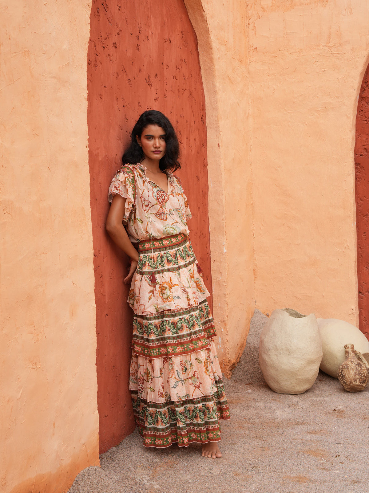 A woman stands barefoot in a rustic outdoor setting with textured orange walls, wearing a long tiered bohemian maxi dress with a floral and geometric print. Large clay pots sit on the ground to her right.