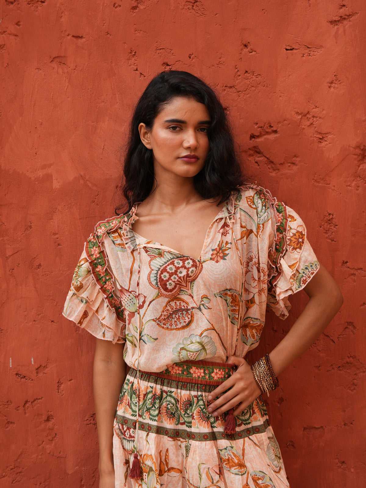 A woman wearing a bohemian-style floral print dress with a matching belt, standing against a textured terracotta wall.