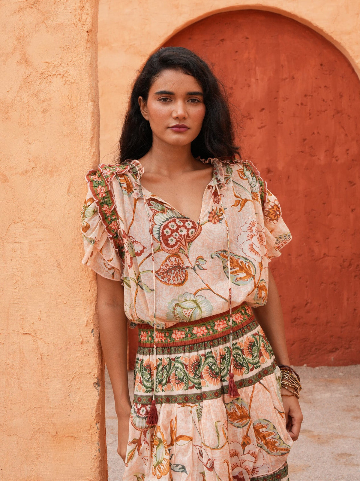 A woman wearing a bohemian-style printed dress with puff sleeves and a patterned belt, standing in front of a textured orange stucco wall with an archway.
