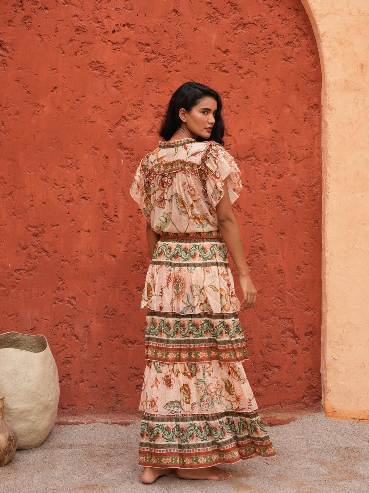 A woman stands in front of a textured terracotta archway wearing a long, tiered bohemian maxi dress with a beige base and colorful floral patterns. The scene is set in a warm, rustic outdoor environment with earthy clay pots on the ground.