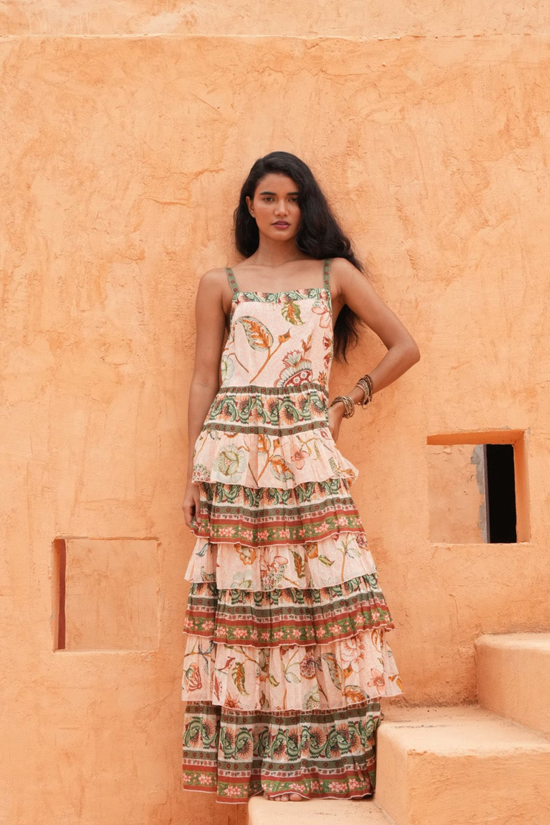 A woman stands on outdoor stone steps in front of a textured orange adobe wall, wearing a sleeveless tiered maxi dress with a cream base and colorful botanical and geometric patterns.