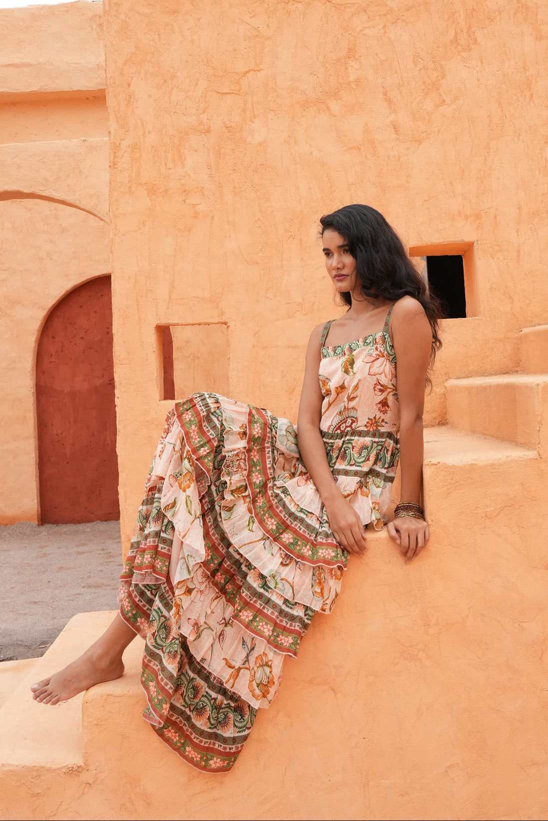 A woman sits barefoot on terracotta-colored stone steps in front of a matching stucco building, wearing a sleeveless, patterned maxi dress with a cream base and green, orange, and red motifs.