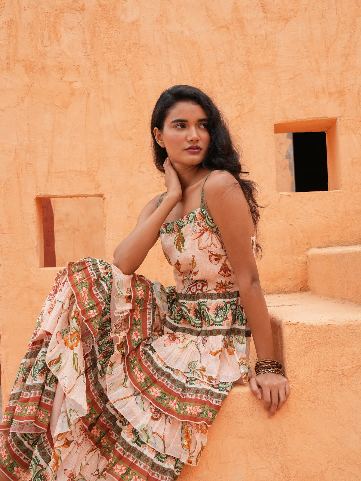 A woman wearing a sleeveless ruffled maxi dress with a colorful floral and geometric print, sitting on terracotta-colored stone steps against a textured stucco wall.