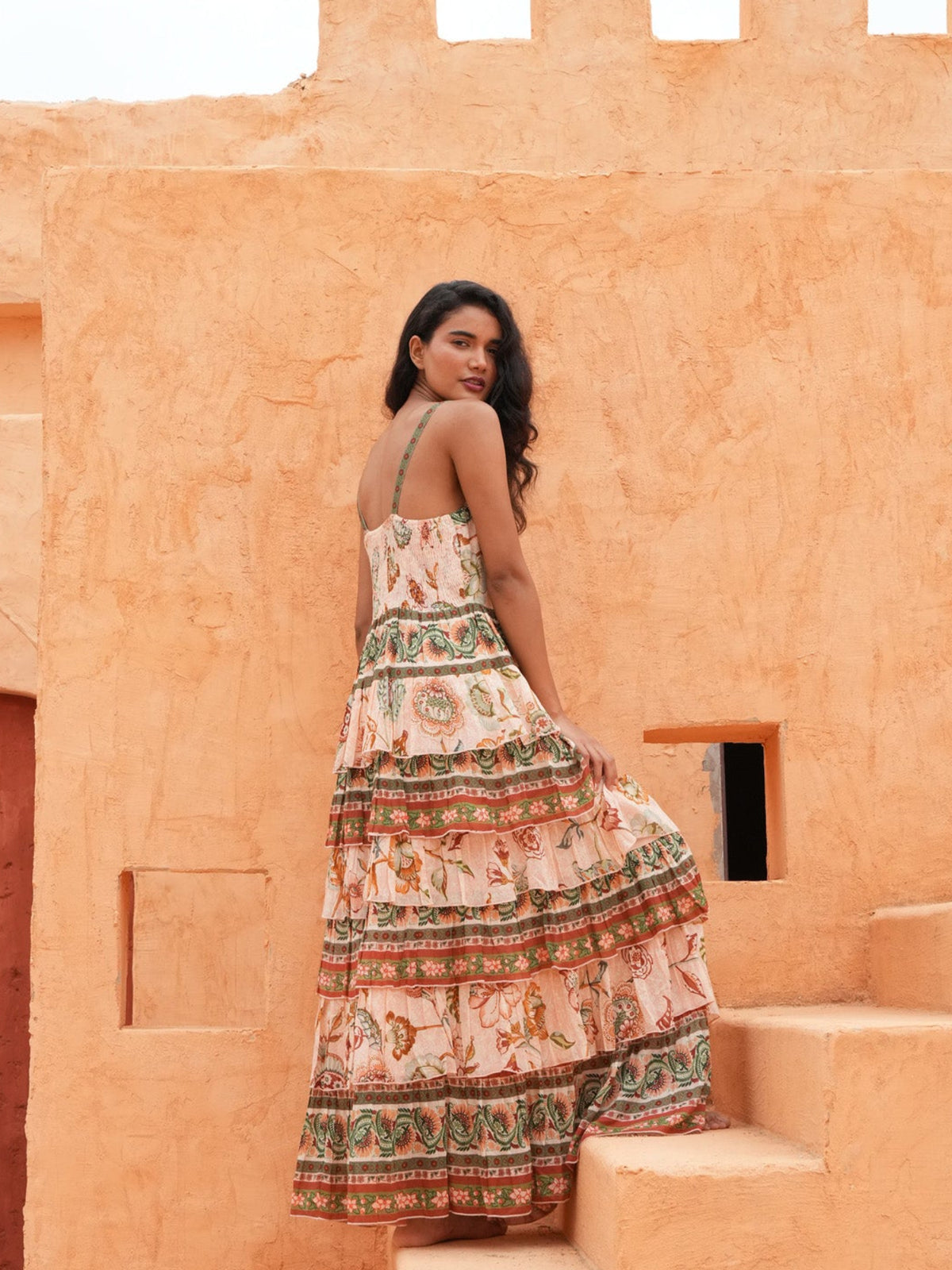 A woman wearing a tiered bohemian maxi dress with a colorful animal print stands on the steps of a textured orange clay building, looking back over her shoulder.