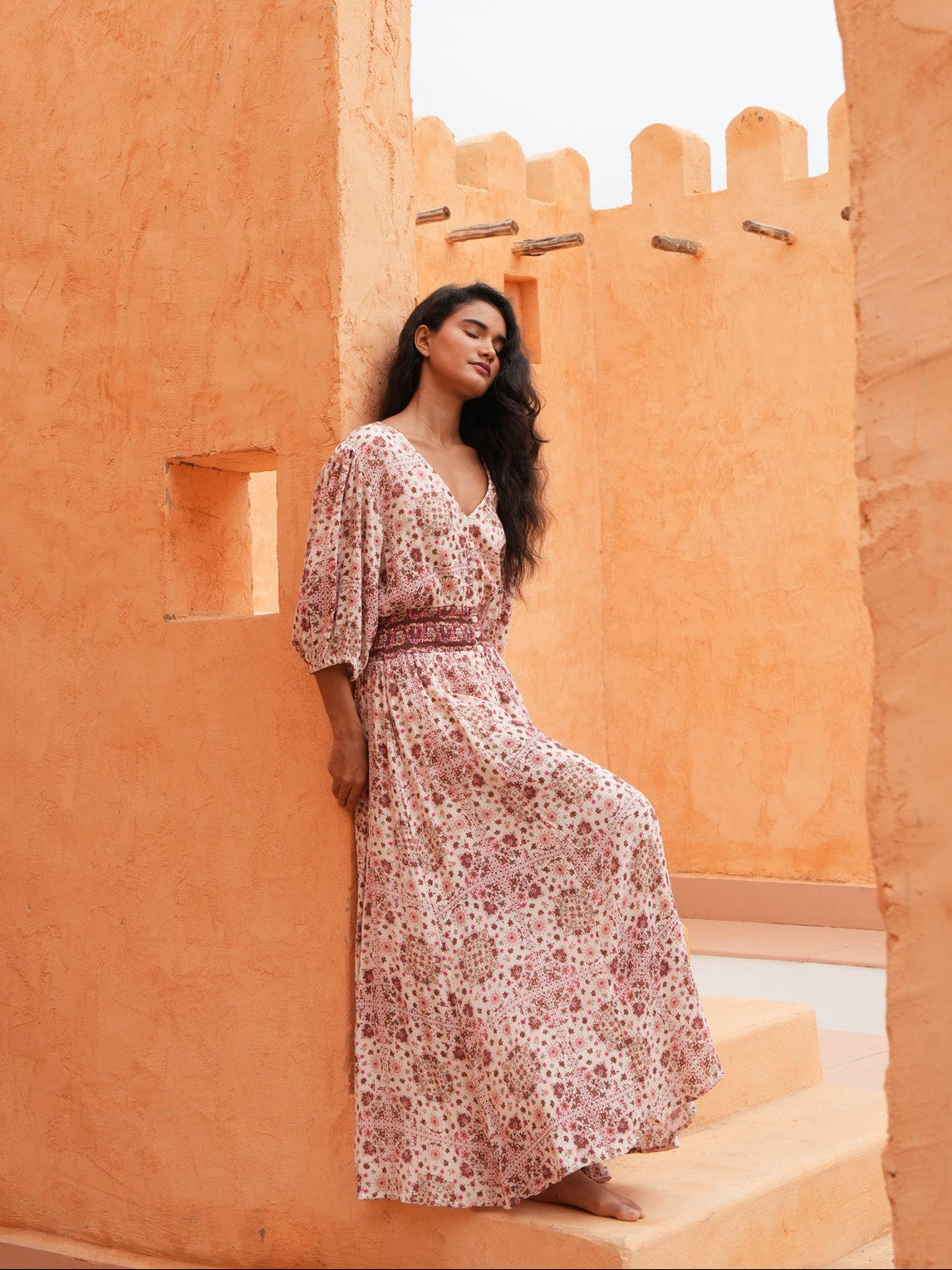 A woman wearing a long-sleeved, knee-length floral maxi dress with a matching waist belt, posing on terracotta-colored stucco steps in front of a matching stucco wall.