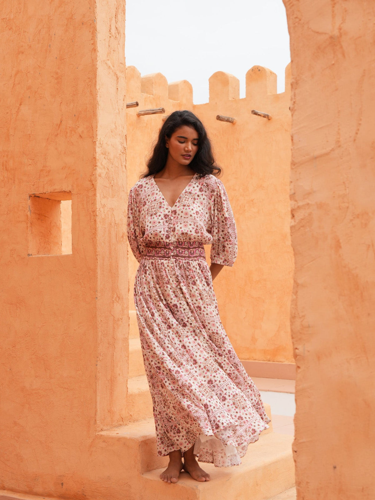 A woman wearing a long, loose floral maxi dress stands barefoot between warm orange stucco walls in an outdoor architectural setting.