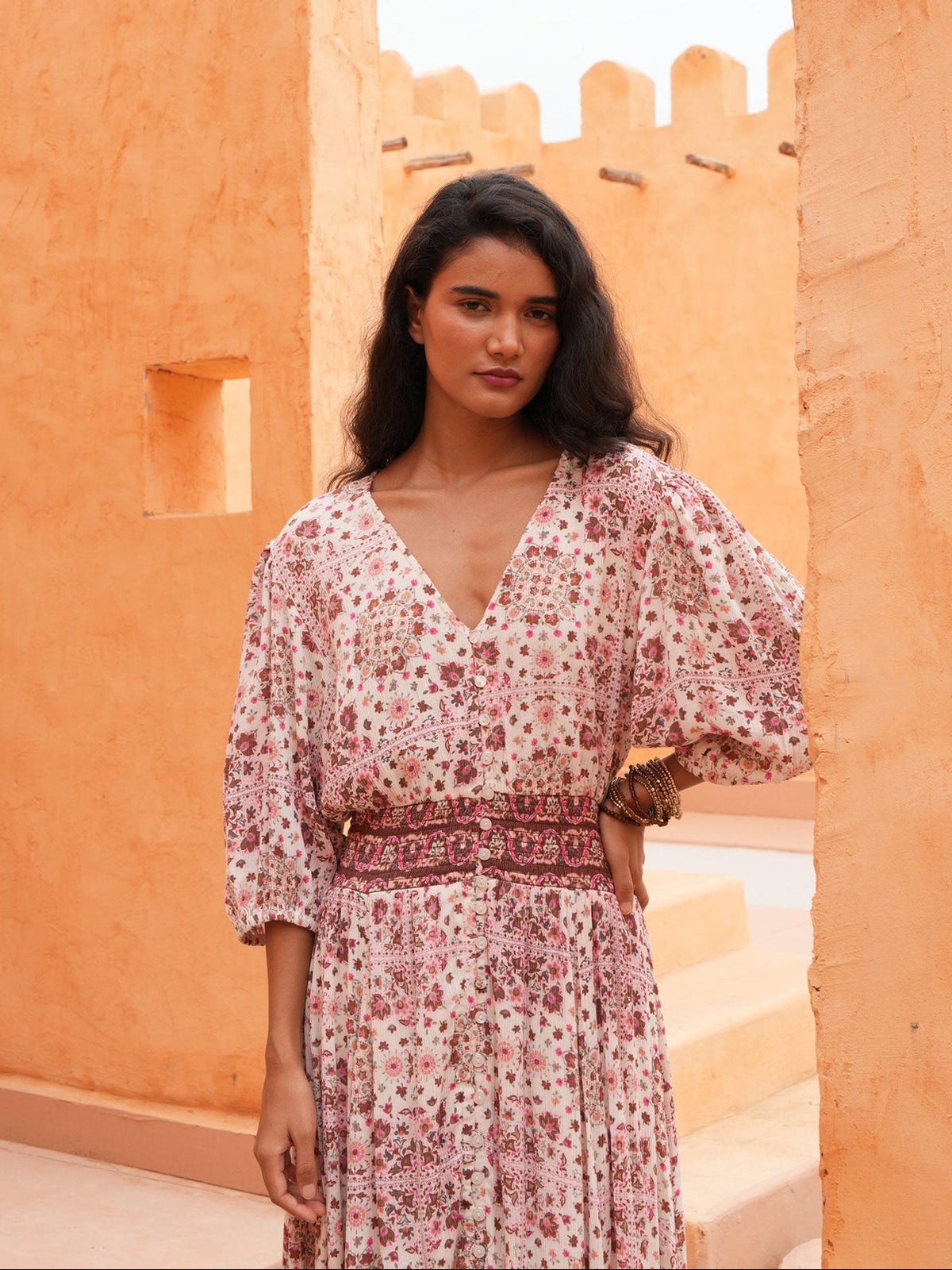 A woman wearing a pink and white floral print maxi dress with puff sleeves and a matching belt, standing in a desert courtyard with orange stucco walls.