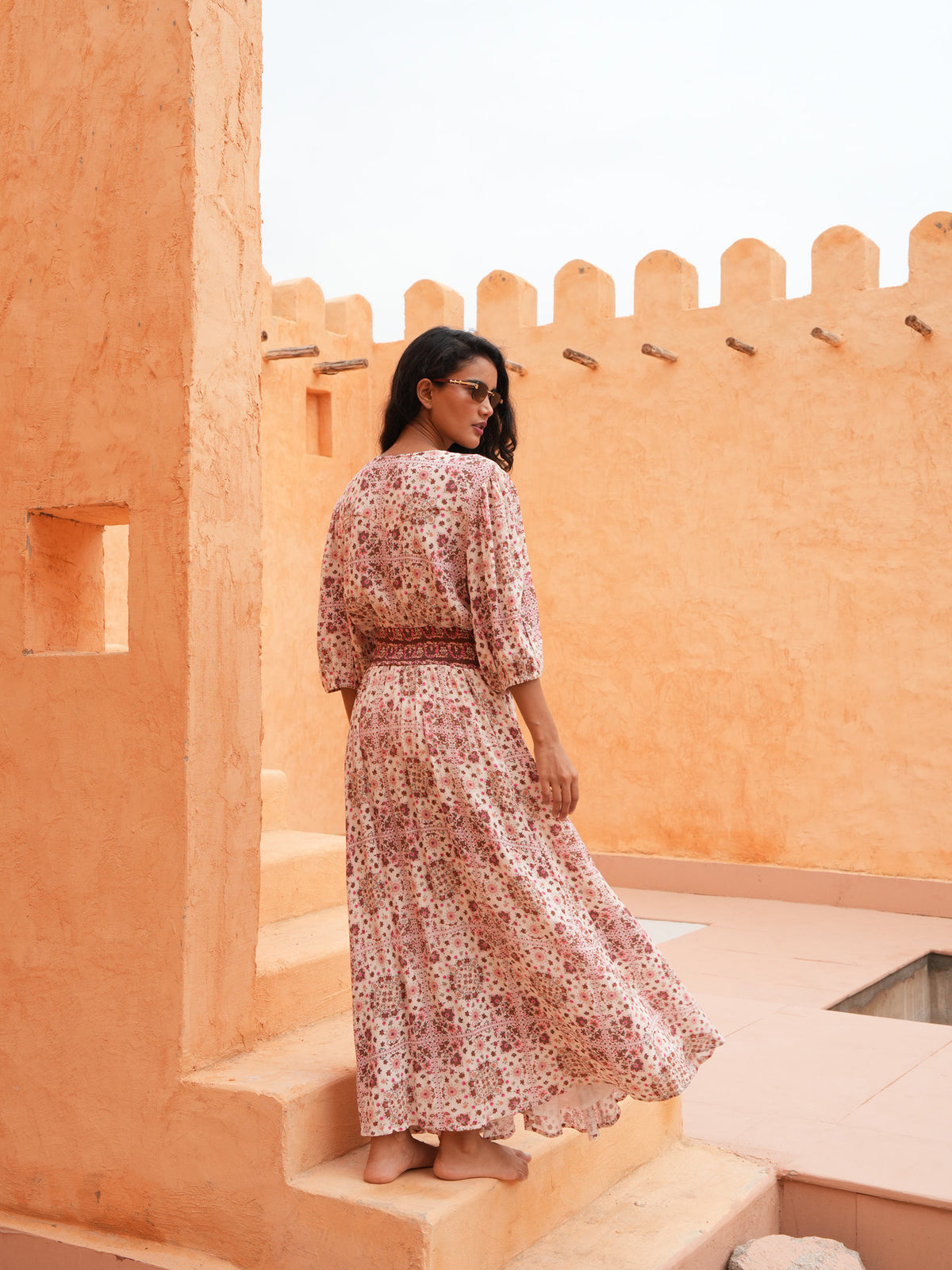 A woman wearing a long-sleeved, knee-length floral dress with a matching waist belt, standing barefoot on stone steps in front of a warm orange stucco wall.