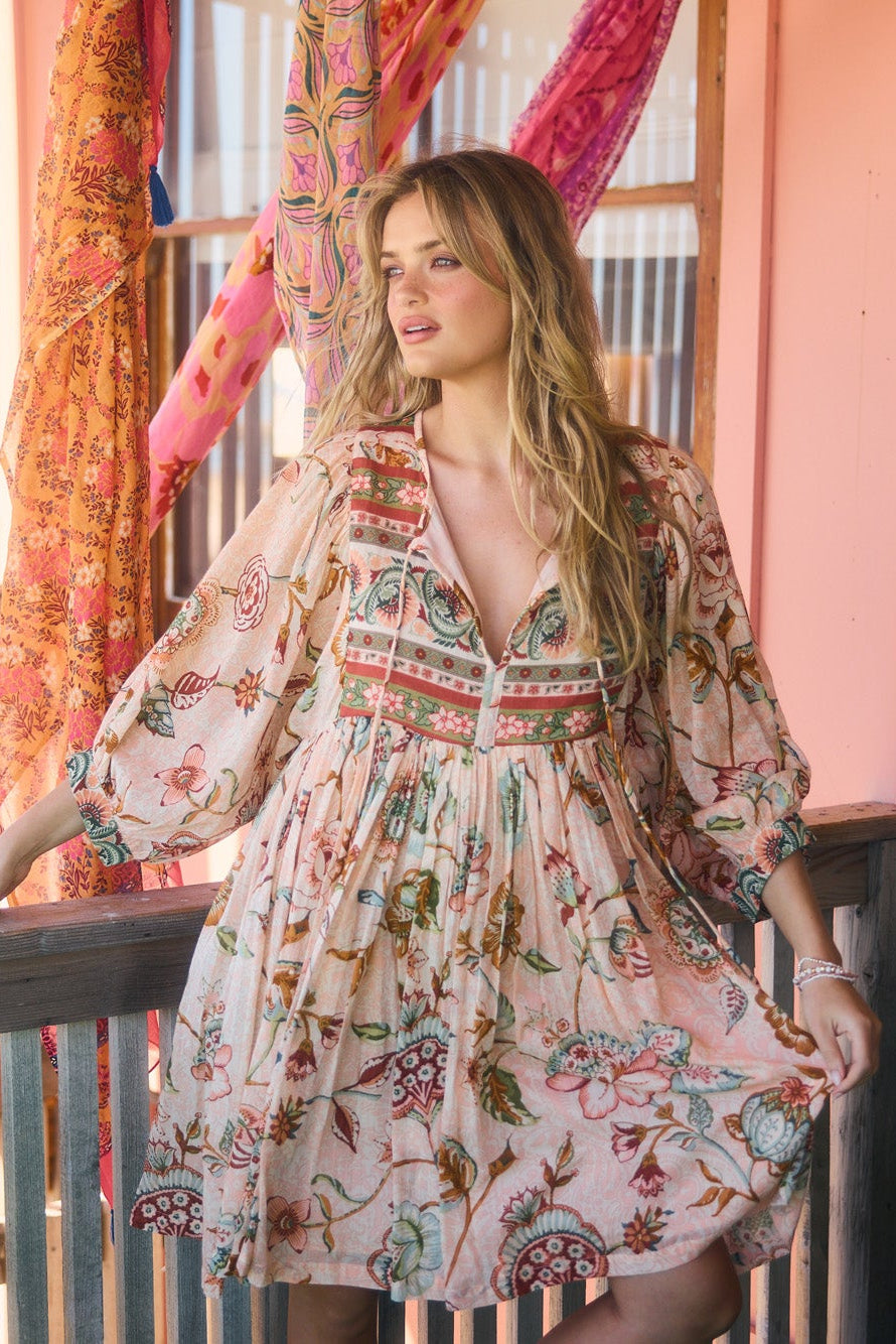 A woman wearing a bohemian-style floral print dress leans on a wooden railing on a porch. The dress has a light pink base with red, green, and blue patterns.
