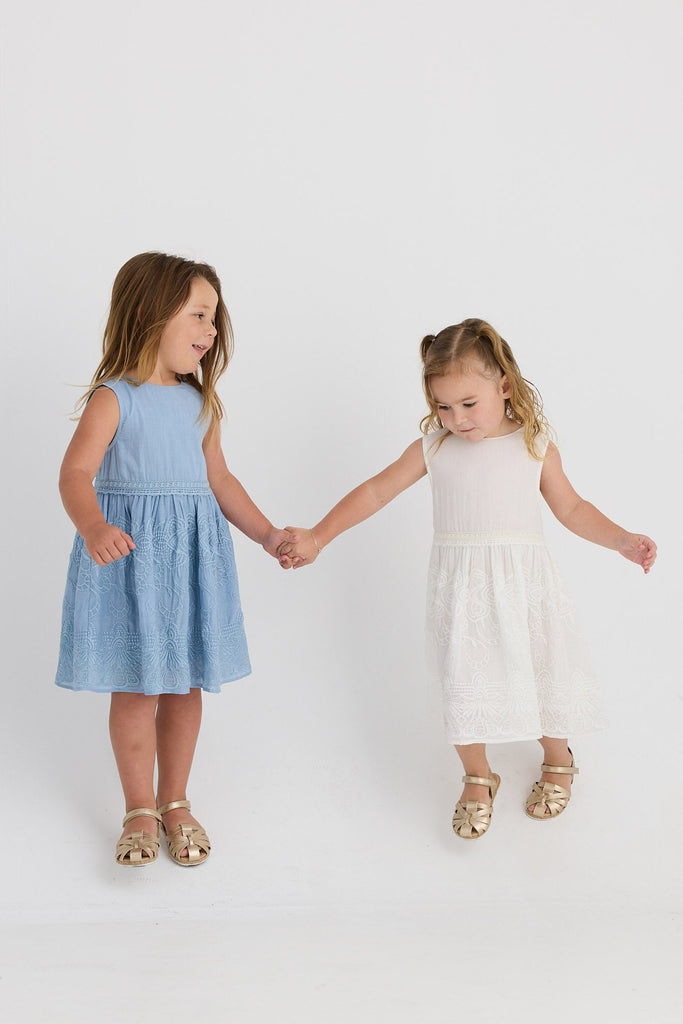 Two young girls holding hands, one in a blue dress and the other in a white dress, on a plain background.