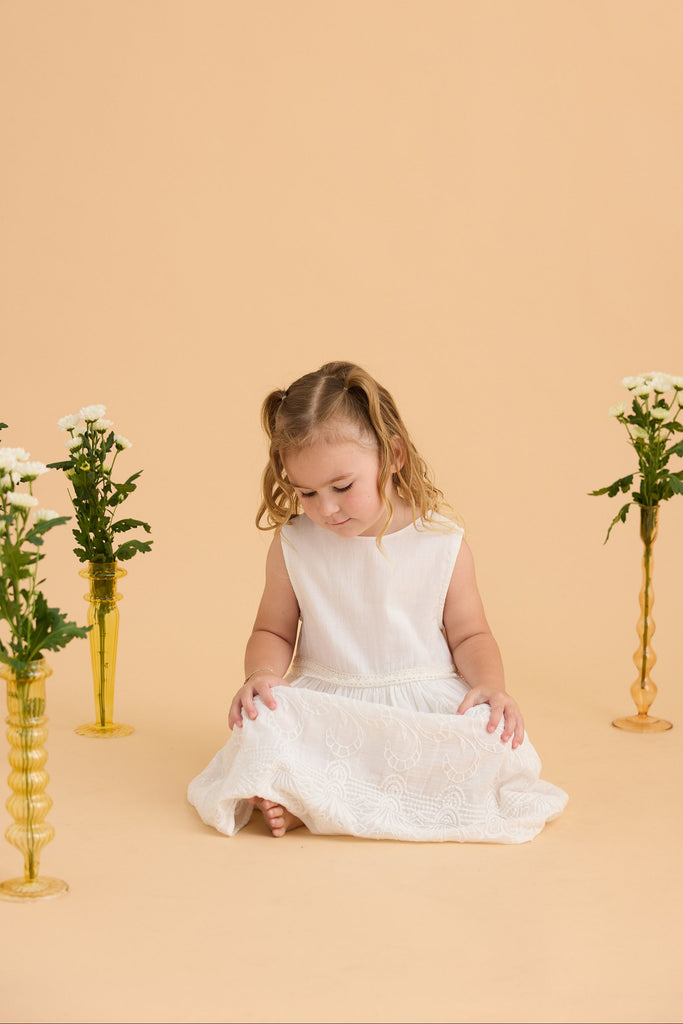 Young girl in a white dress sitting on a beige background with floral arrangements.