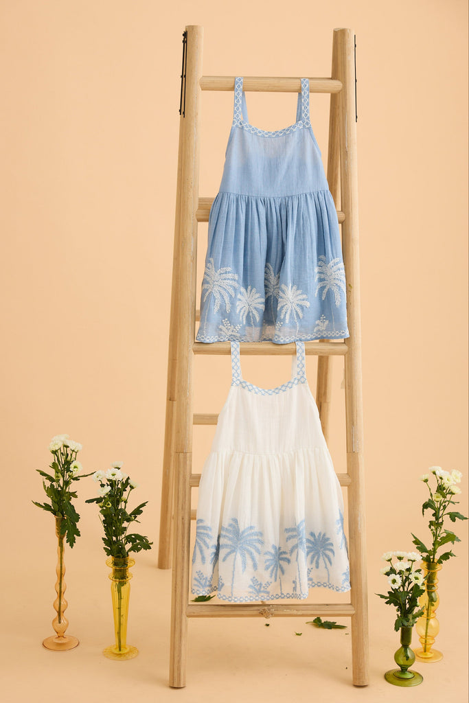 Two dresses, one blue and one white, on a wooden ladder with decorative plants on a beige background.