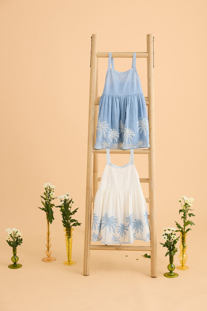 Two dresses, one blue and one white, on a wooden ladder against a beige background with decorative plants.