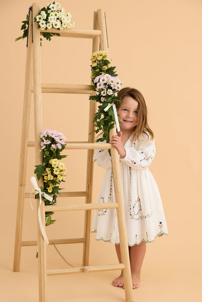 Young girl in a white dress standing next to a wooden ladder with floral decorations on a beige background