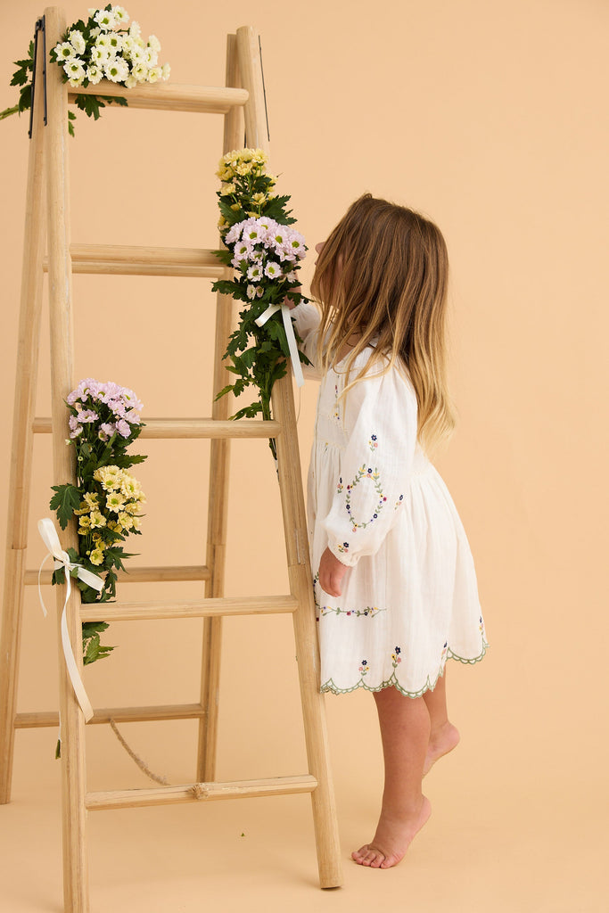 Child in a white dress standing next to a ladder with floral arrangements on a beige background